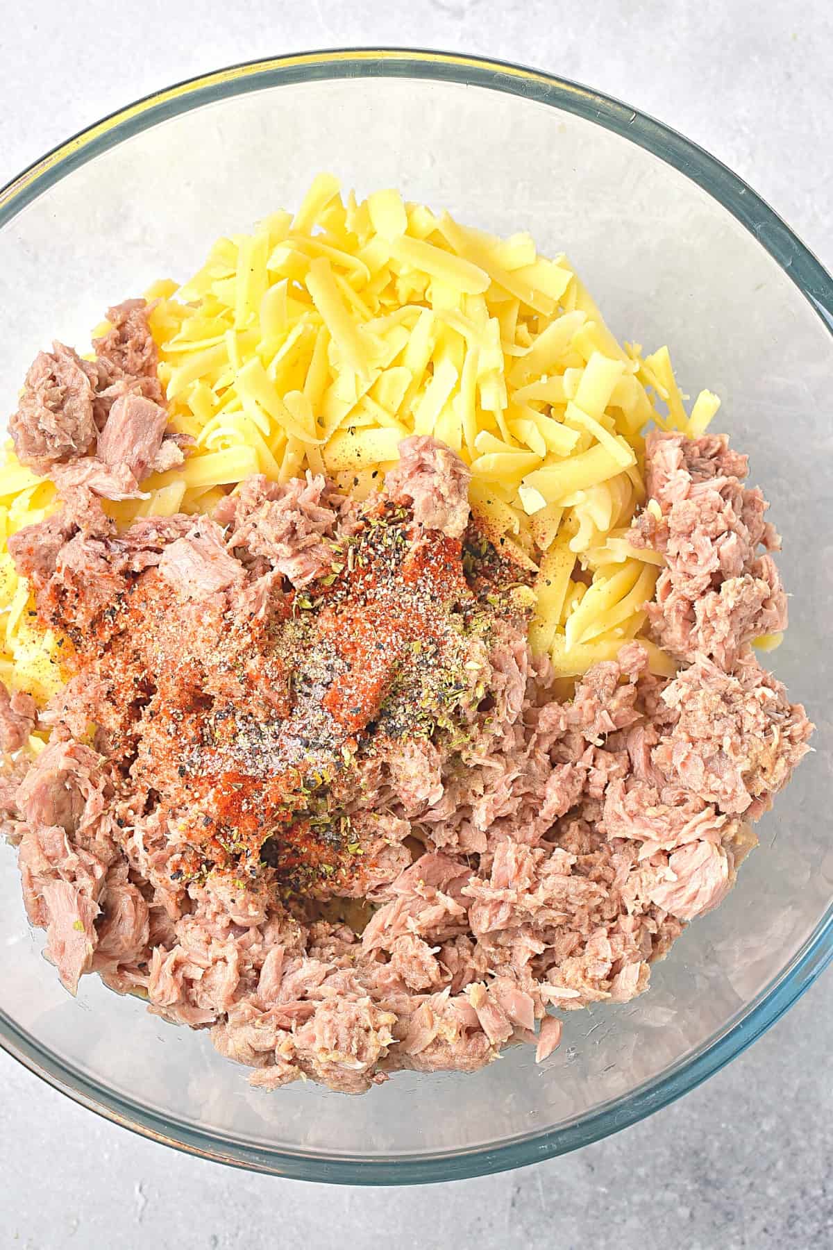 Canned tuna, shredded cheese and spices in a glass mixing bowl.