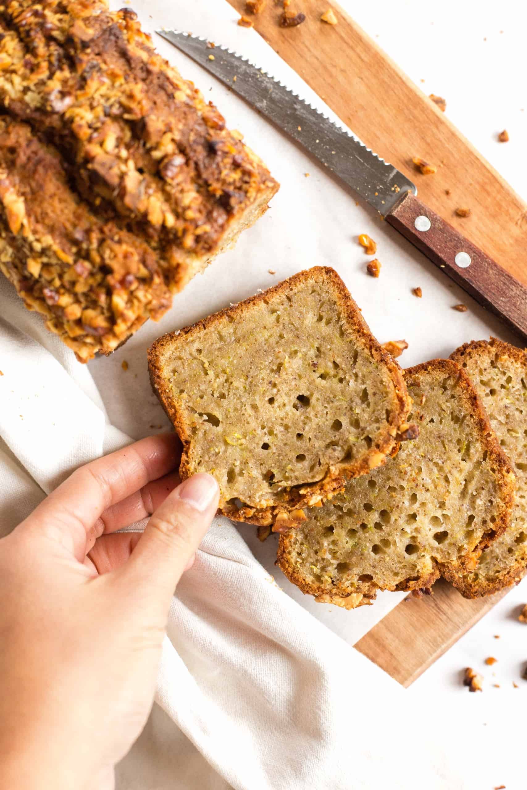 Reaching for a slice of gluten-free zucchini bread from a wooden board.