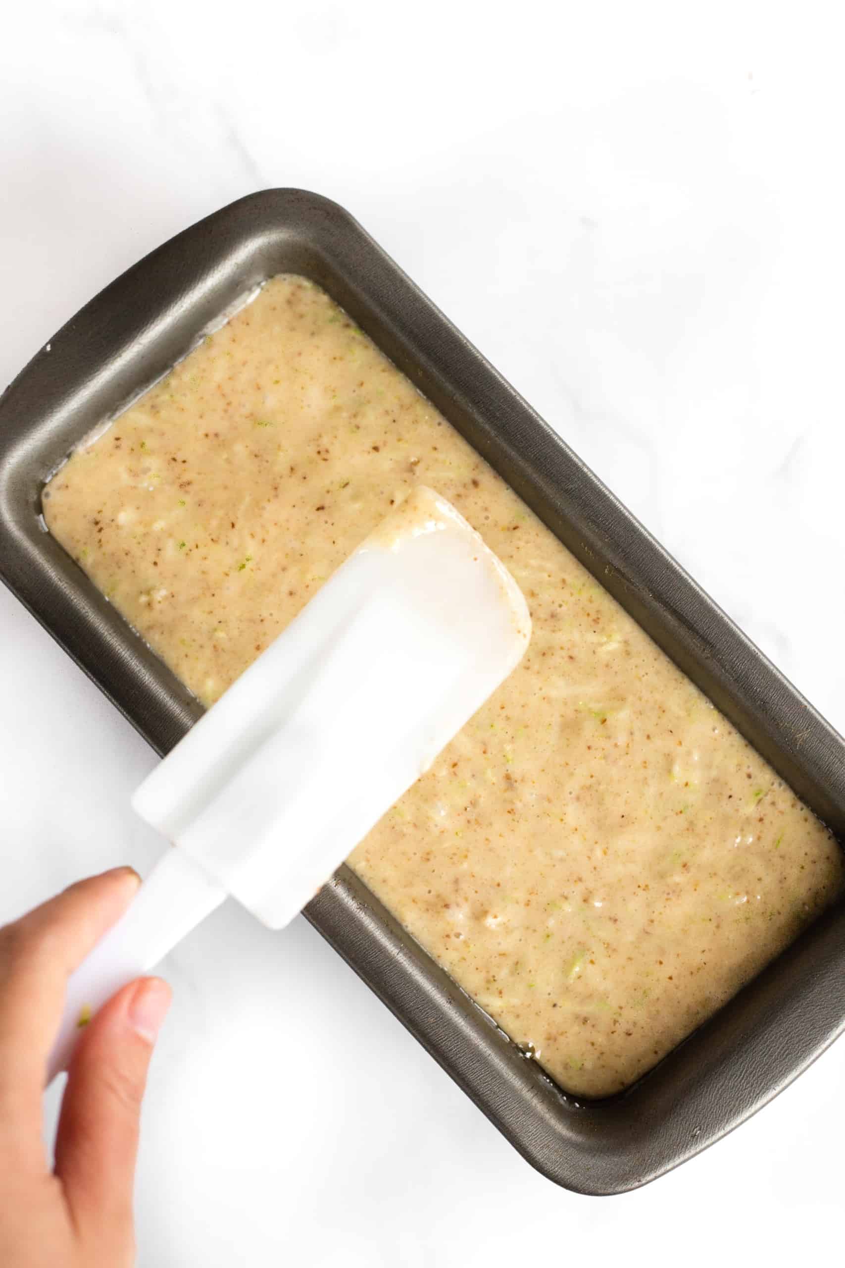 Using a spatula to smooth the top of batter in a metal loaf pan.