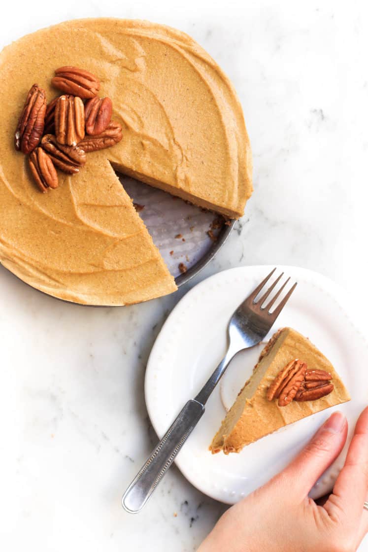 Hand holding a plate with pumpkin cheesecake and a fork alongside the rest of the cake.