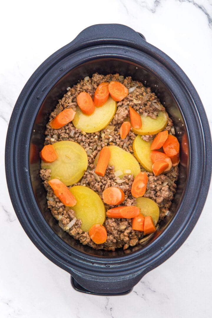 A slow cooker with layered ground beef, potatoes and carrots