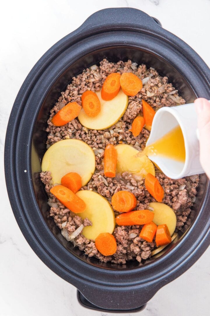 Pouring beef broth into a slow cooker with ground beef, sliced potatoes and carrots.