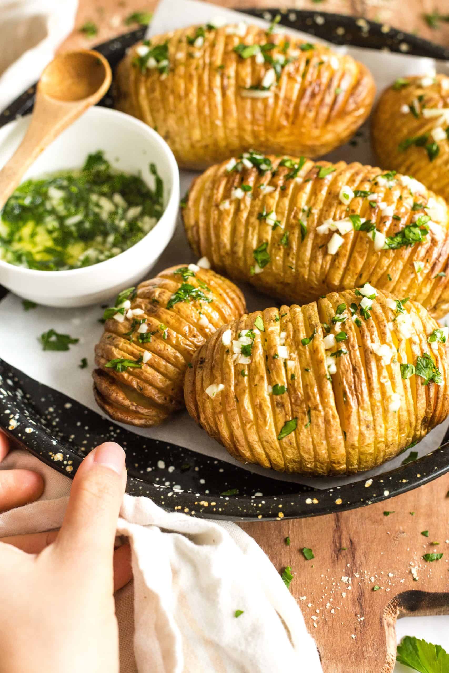 Hand holding a baking sheet with freshly cooked Hasselback potatoes garnished with minced garlic and chopped parsley.