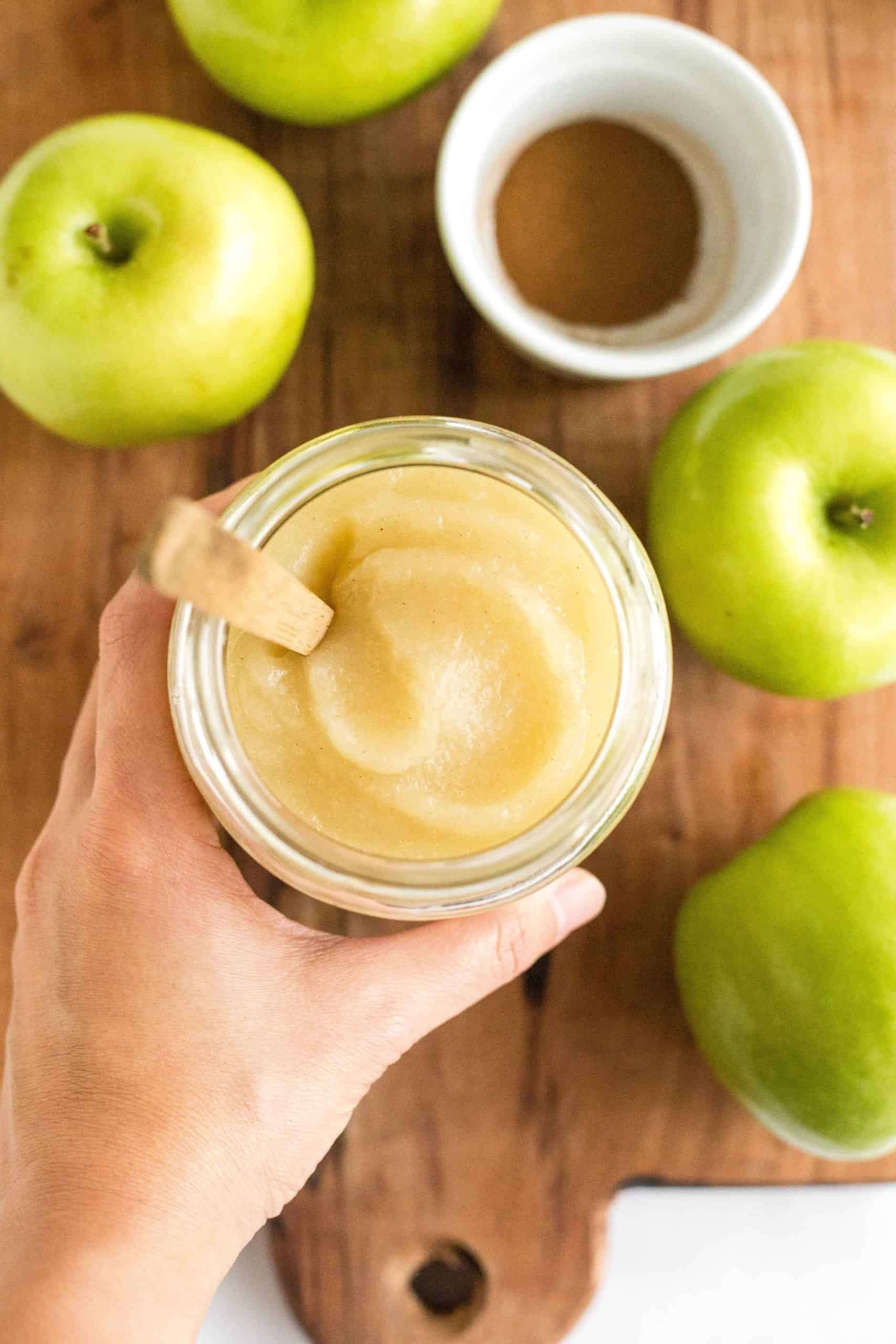 Top view of a hand holding a jar of applesauce with a wooden stick in it. 