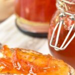Orange preserve spread on sliced bread with jars of marmalade behind.