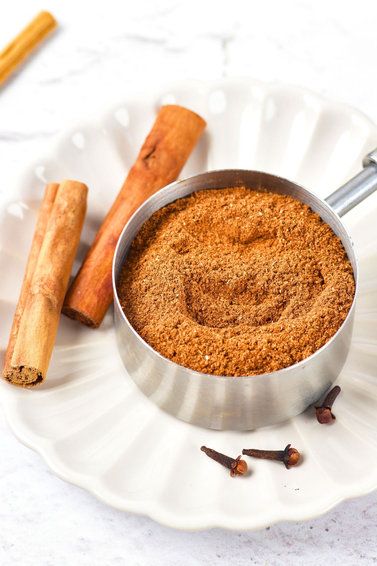 Up close view of a measuring cup with homemade pumpkin spice and whole cinnamon sticks on plate.