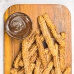 Top down view of churros and a bowl of melted chocolate on wooden board.
