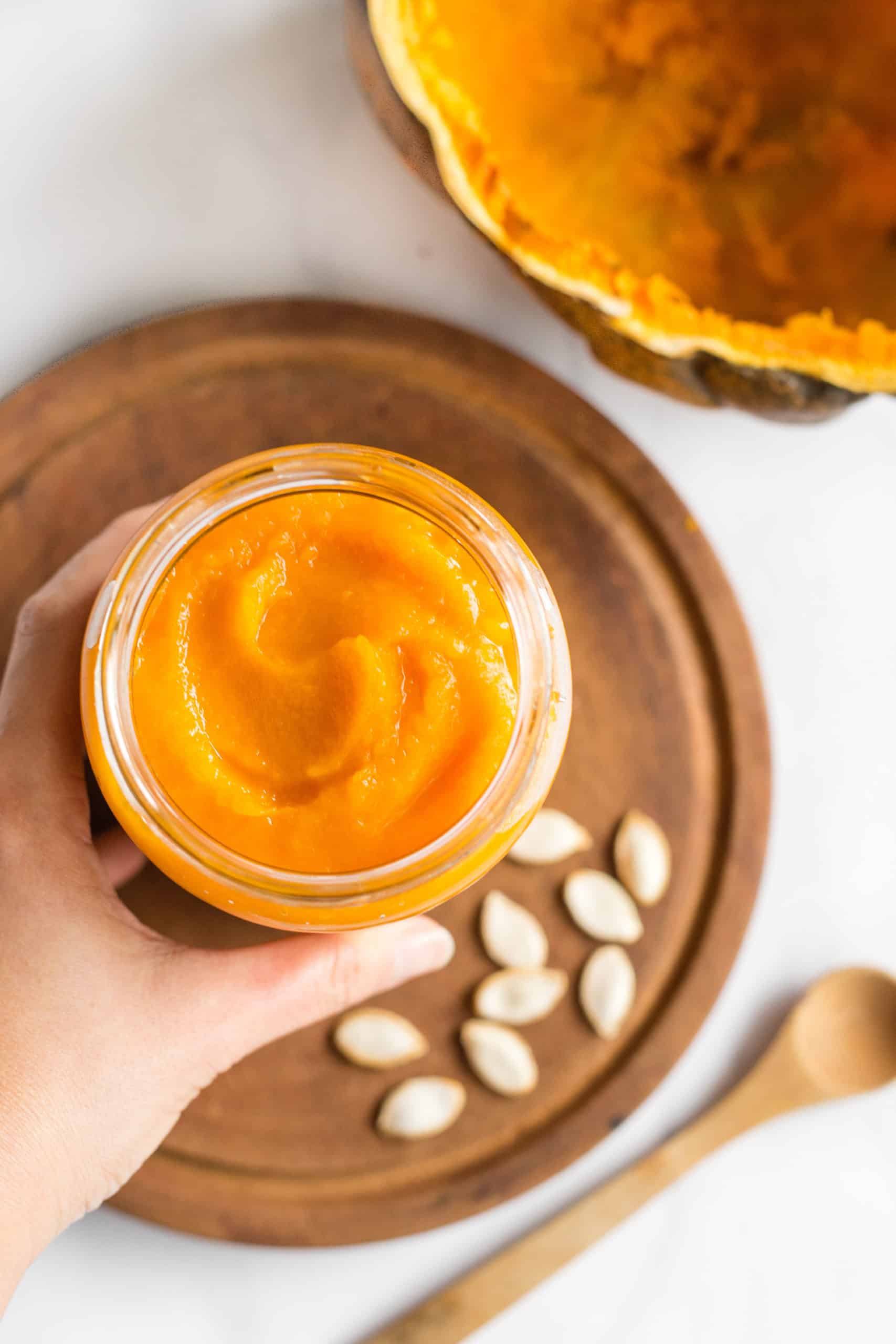 Hand holding a jar of fresh pumpkin puree, and an empty pumpkin shell by the side.