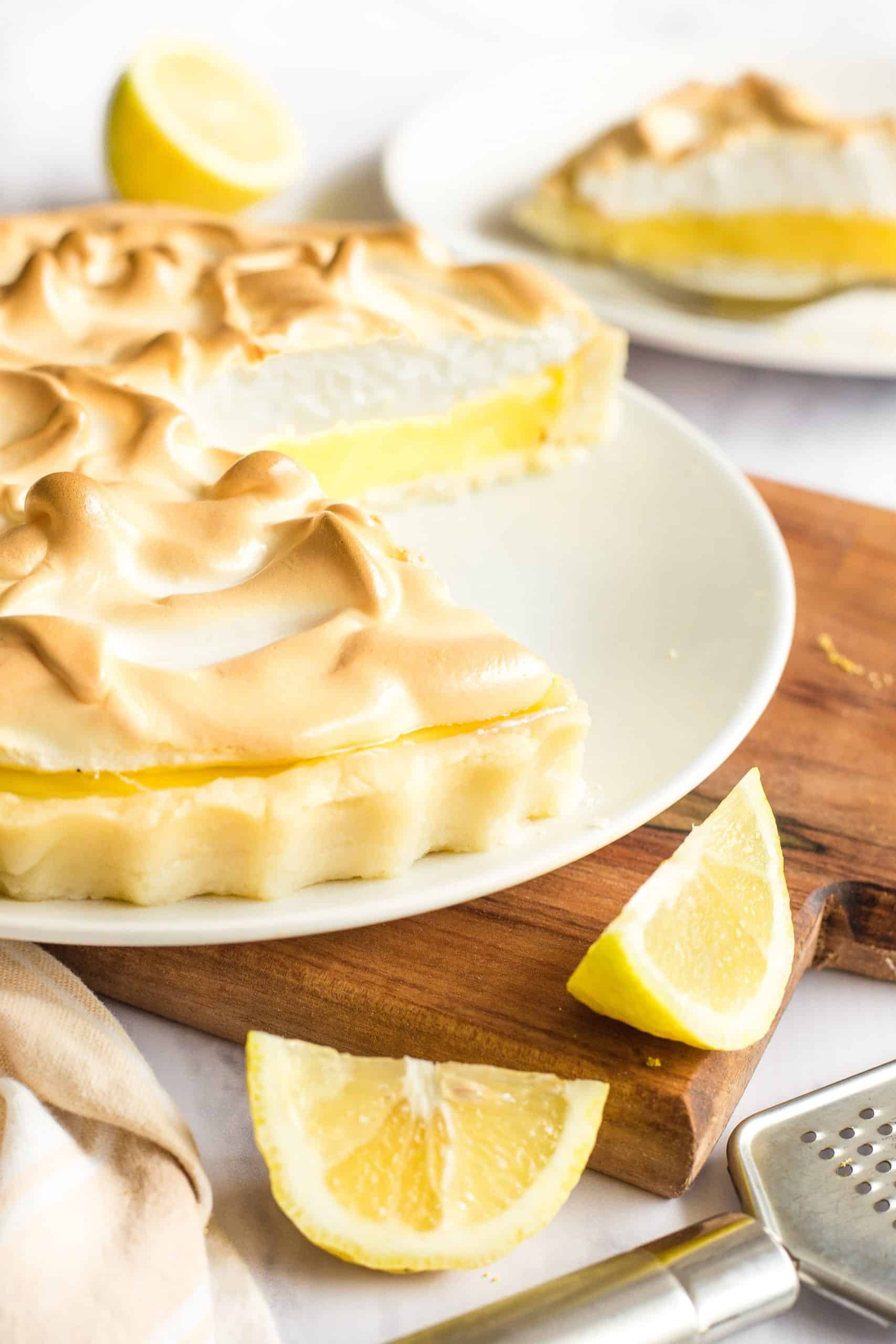 A half-eaten gluten-free lemon meringue pie on a wooden board with cut lemons.