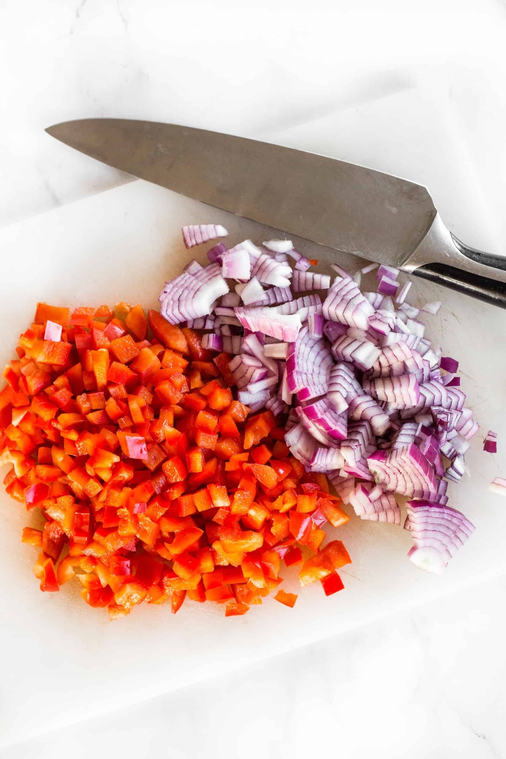 Diced red onions and red bell peppers with large knife on chopping board.