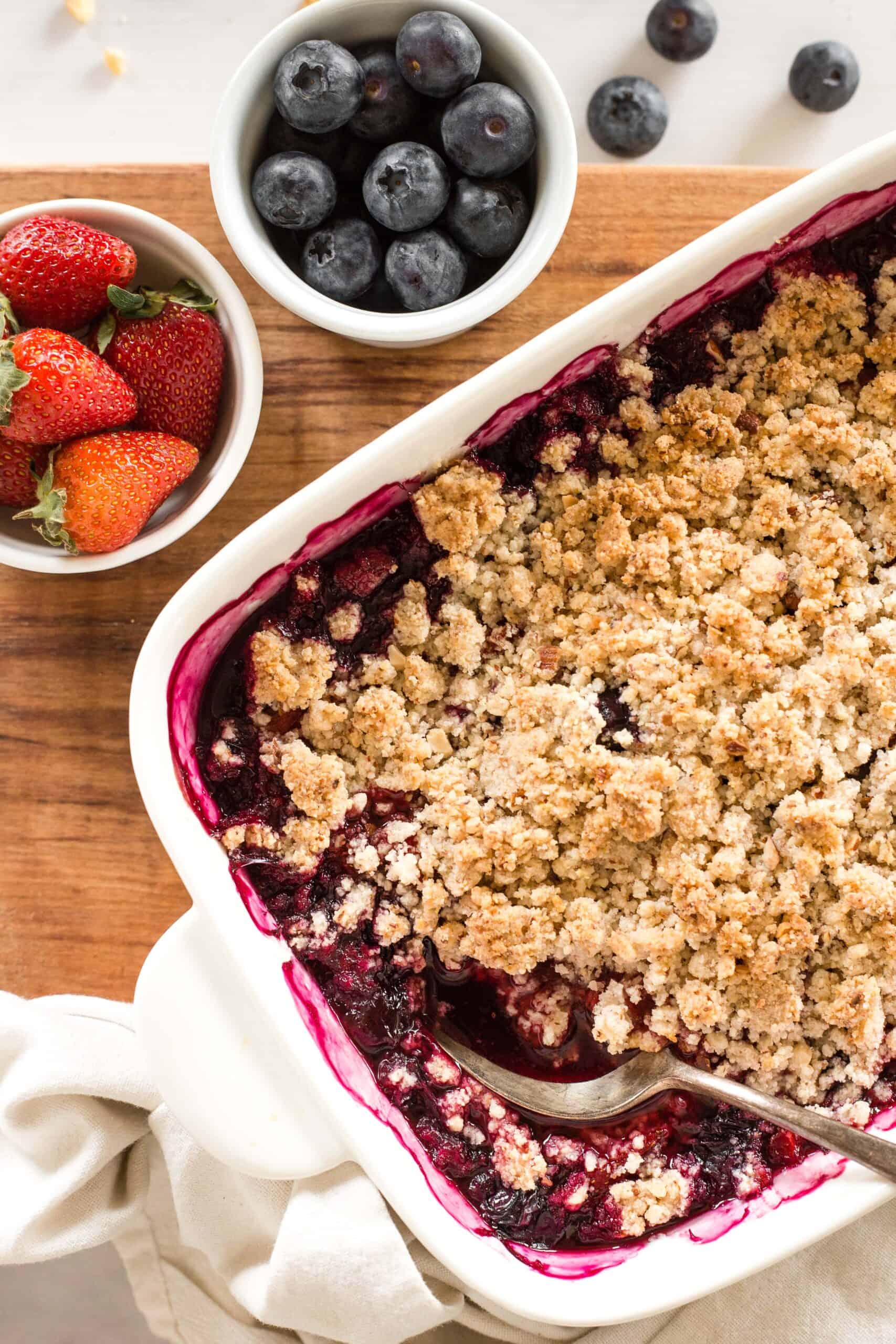 Gluten-free berry crumble and bowls of fresh berries on a wooden board.