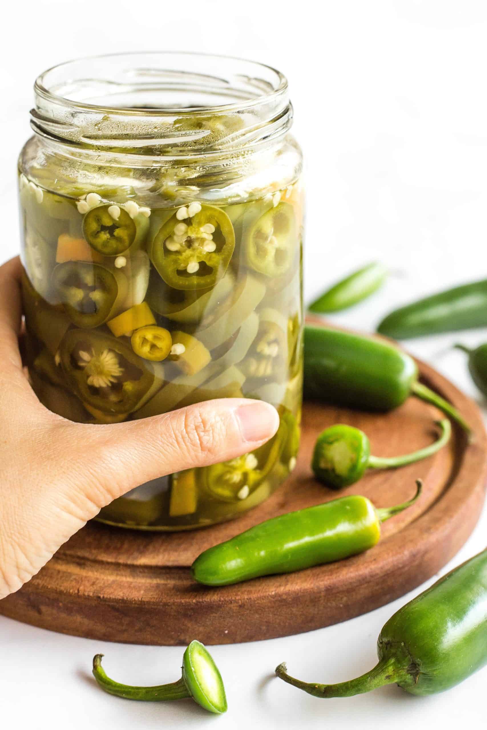 Hand holding an open jar of jalapeño pickles on a wooden board.