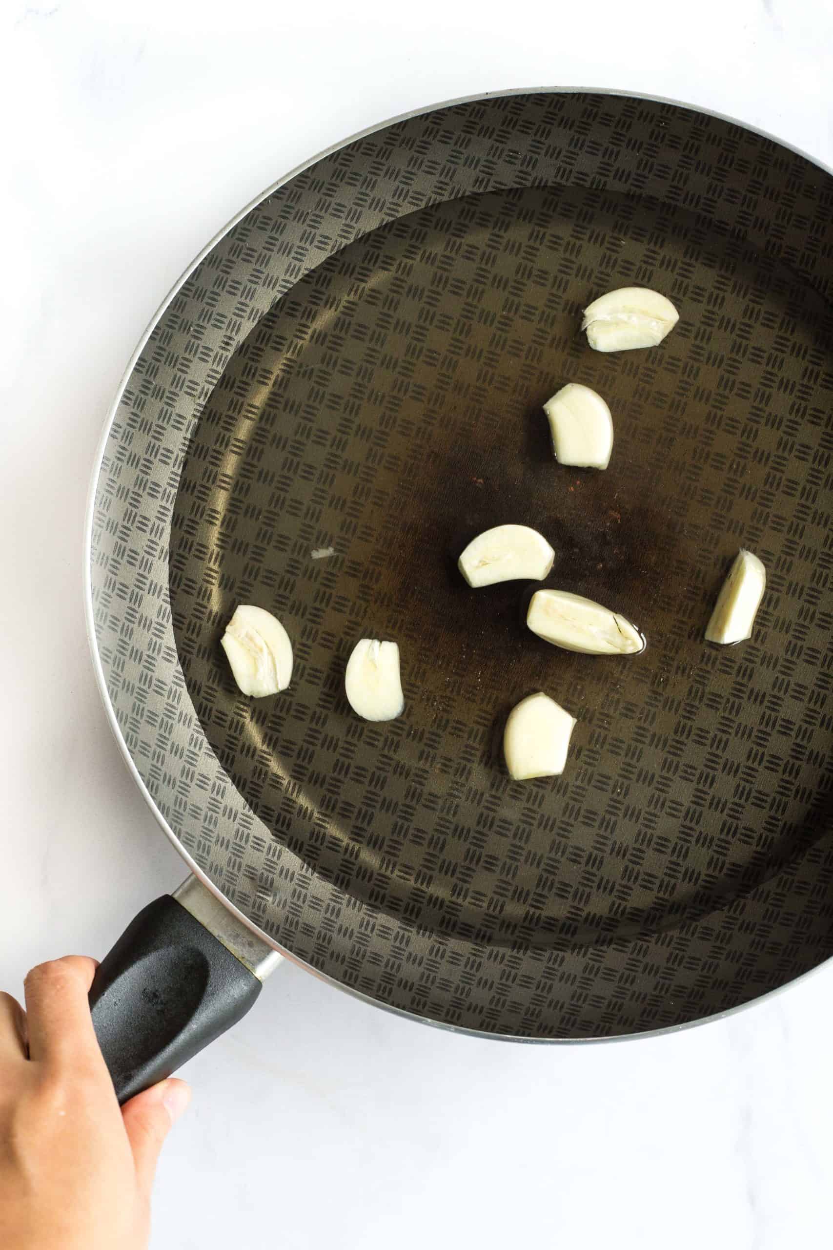 Hand holding a skillet with cloves of garlic in a clear liquid mixture.