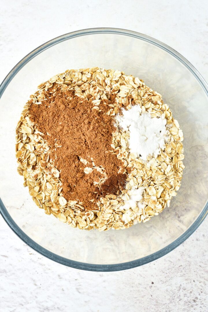 A glass bowl with rolled oats, spices, baking powder and salt.