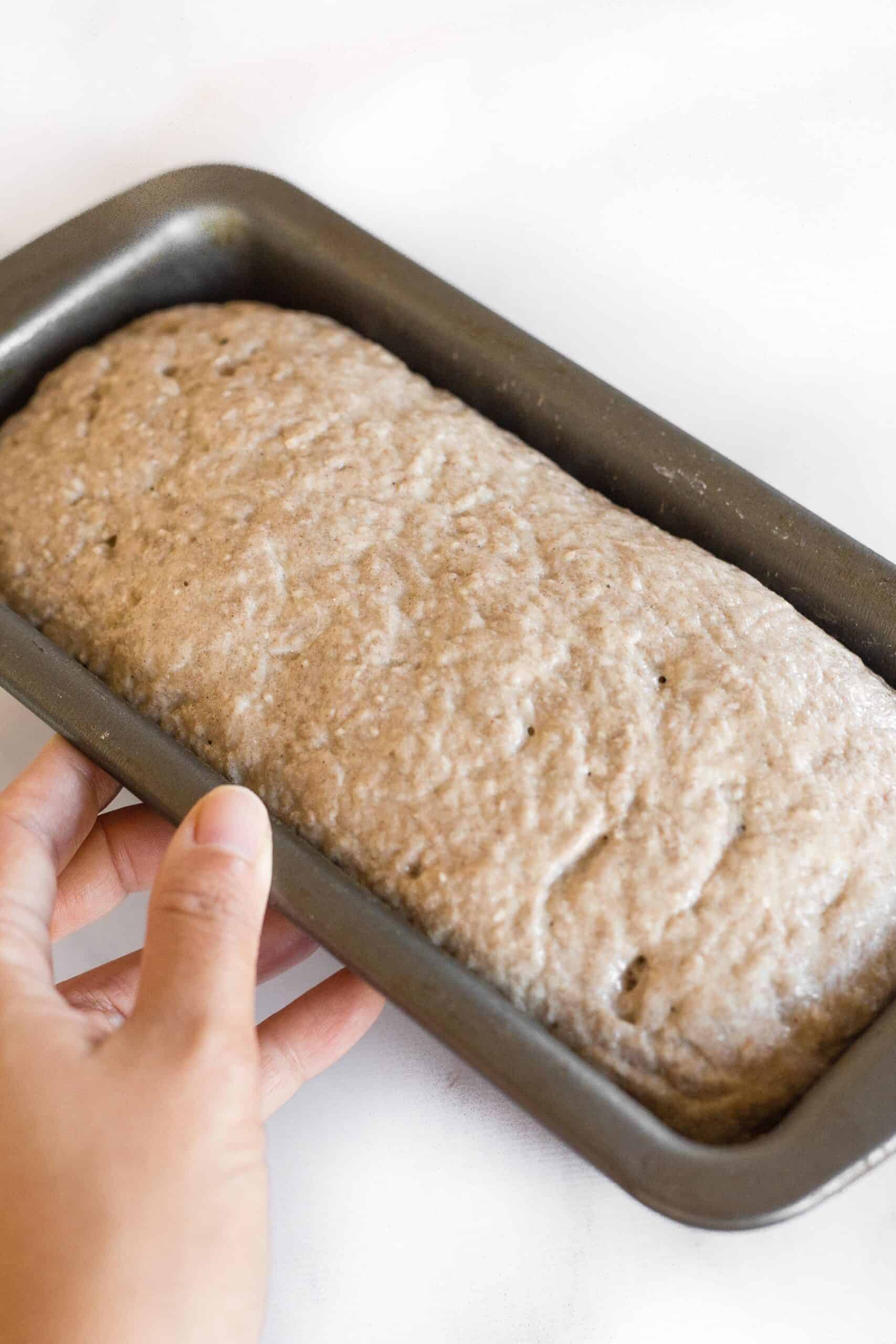 Hand holding a metal loaf pan with risen teff flour bread dough.