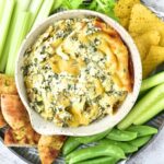 Top down view of a bowl of spinach artichoke dip surrounded with vegetable crudités and chips.