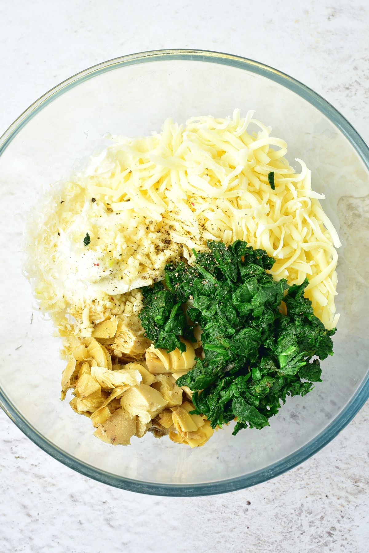 Shredded cheese, wilted spinach, and chopped artichokes in glass bowl