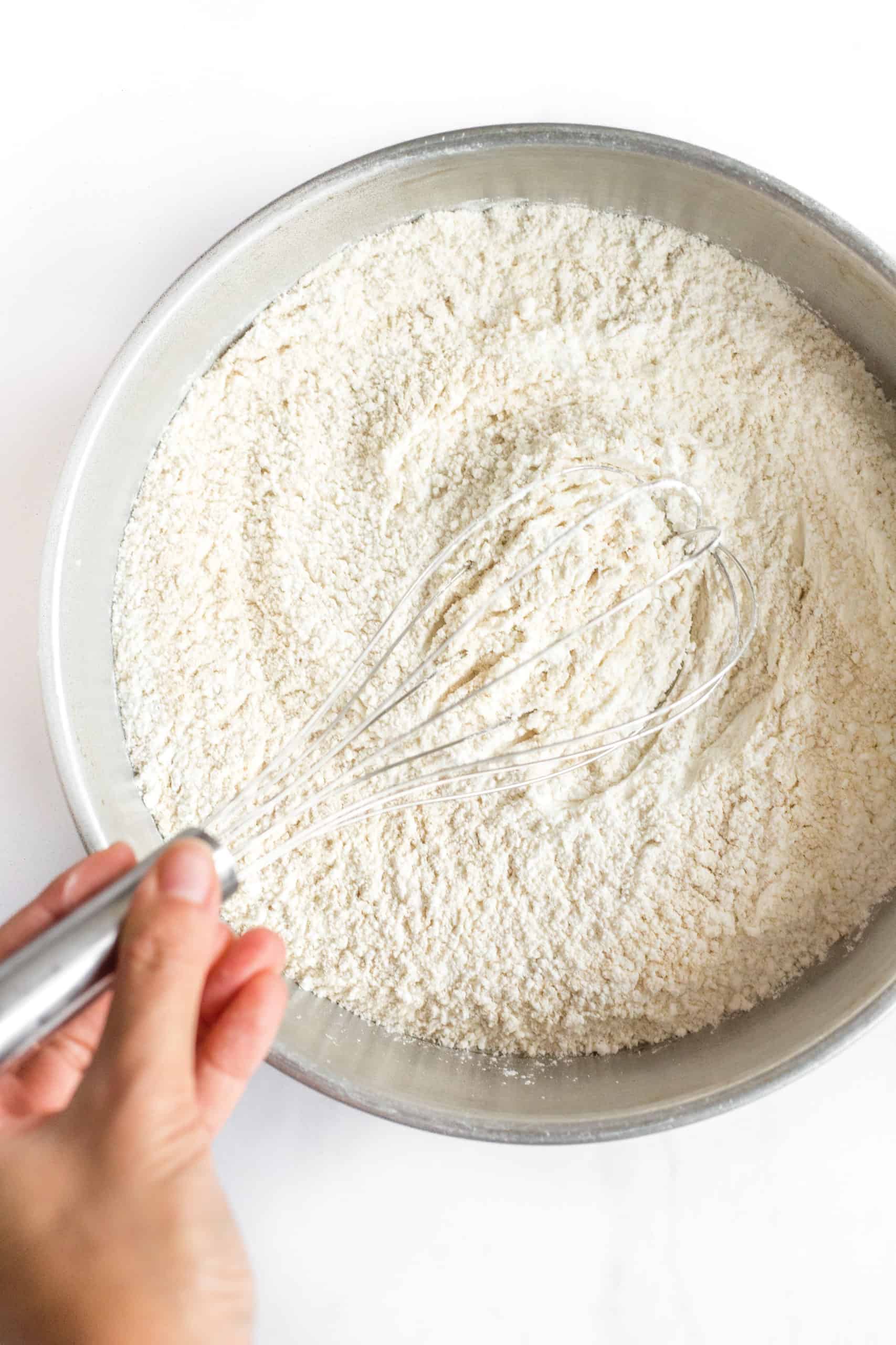 A hand whisking gluten-free all-purpose flour, brown rice flour, and salt together in a large mixing bowl.