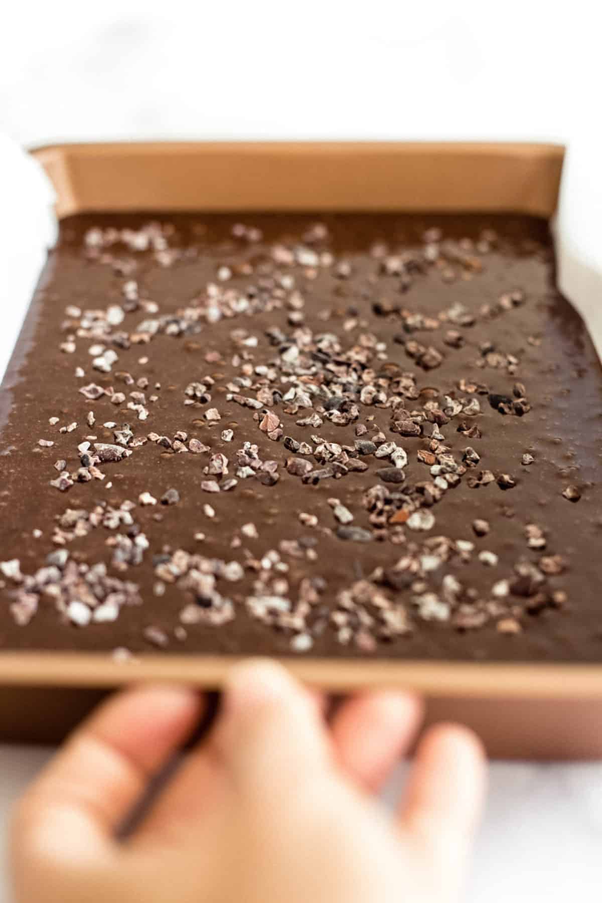 Hand reaching for a pan with gluten-free brownie batter and cacao nibs.