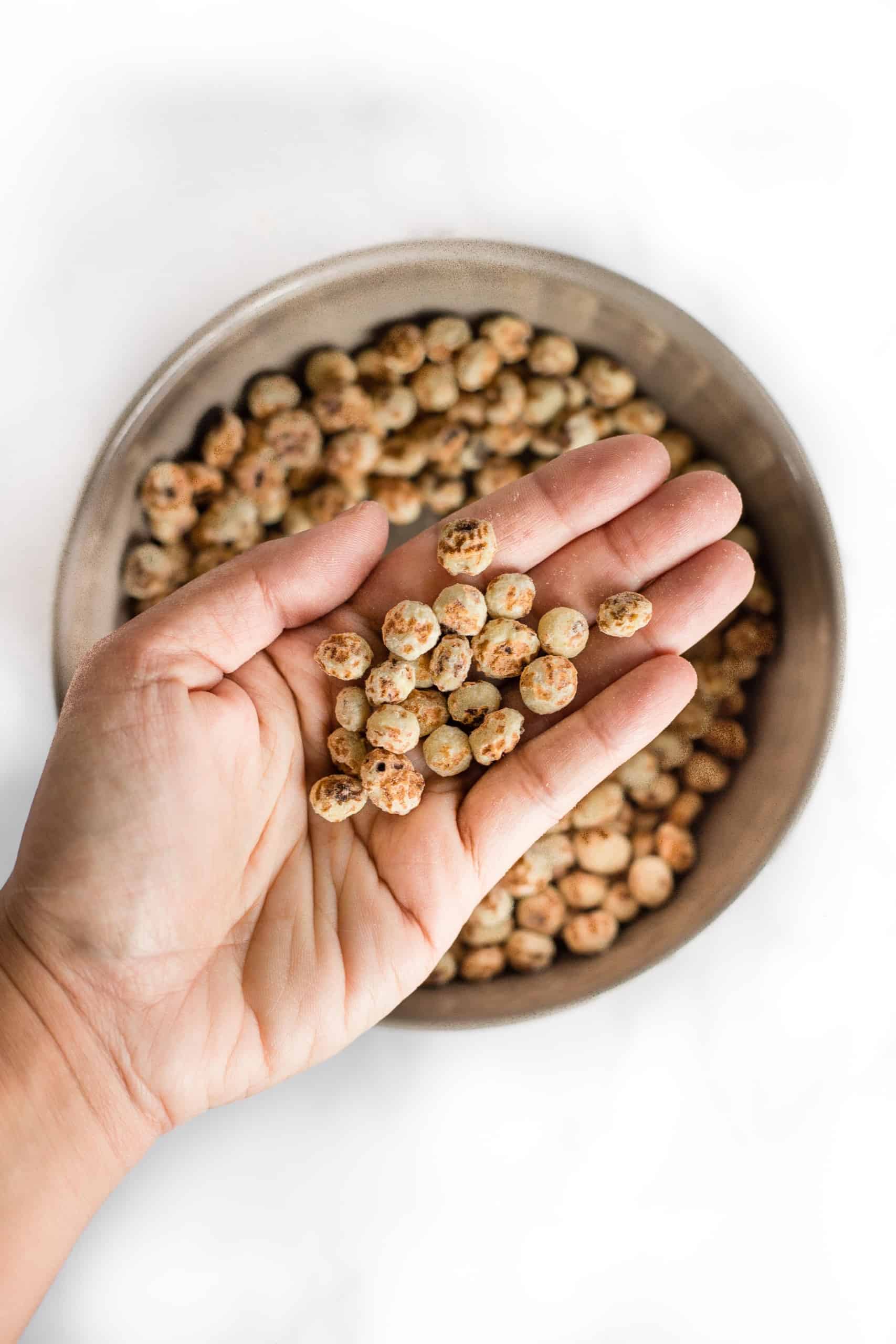 Hand holding up raw, peeled tiger nuts from a bowl.