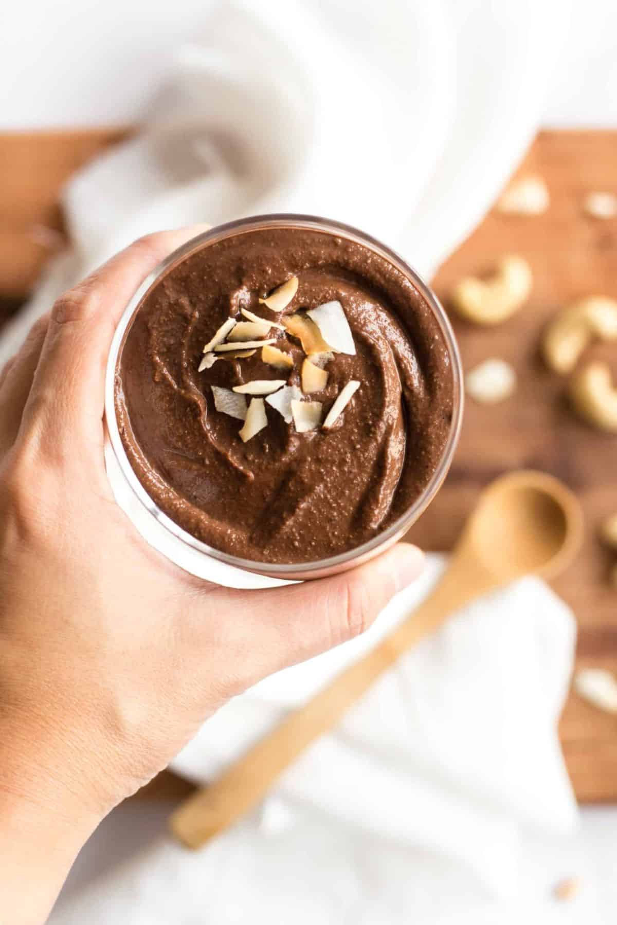 Hand holding a cup of cashew chocolate pudding topped with coconut flakes.