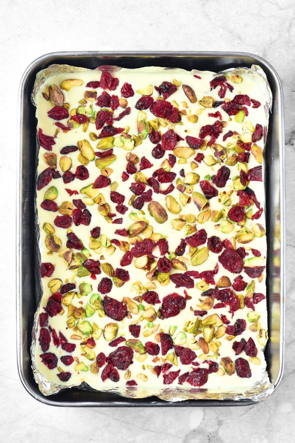 Dried cranberries and pistachios sprinkled over melted white chocolate in baking pan.