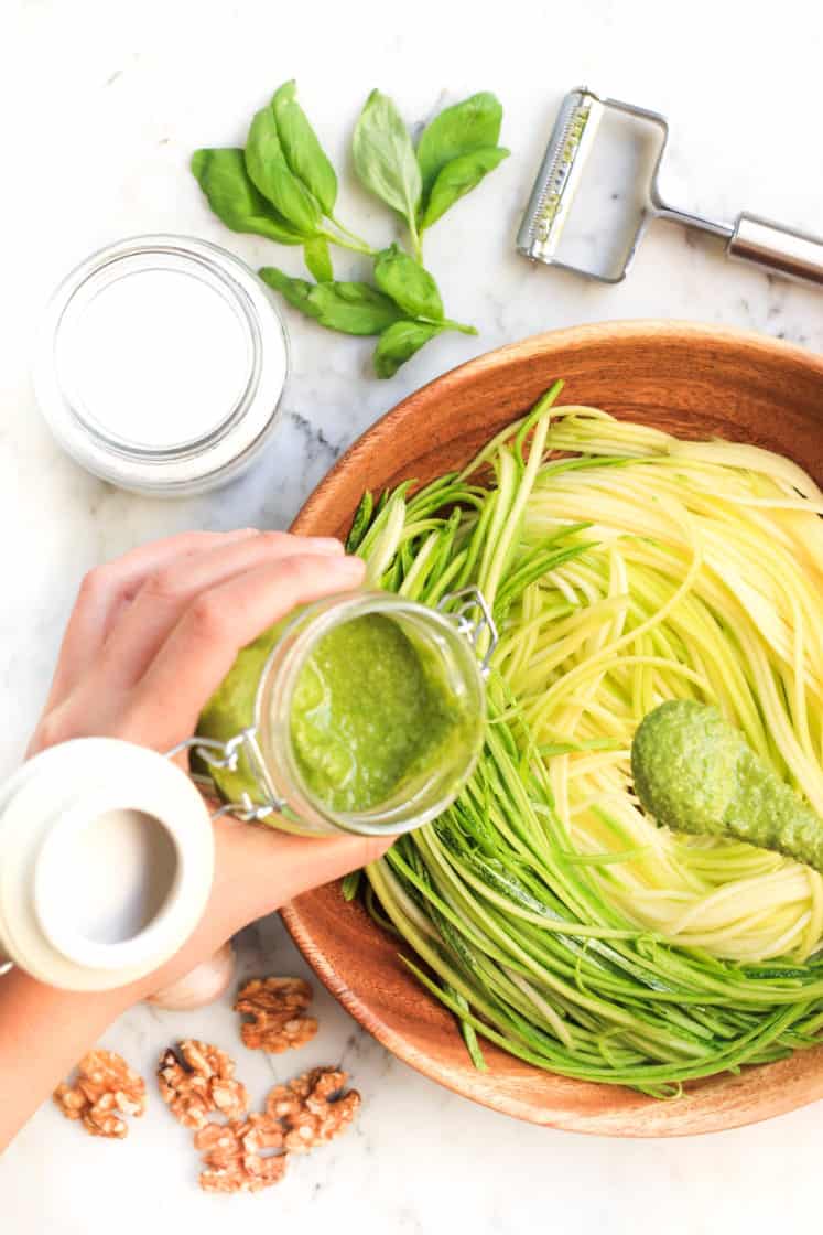 Drizzling pesto sauce over zucchini noodles in a wooden bowl.